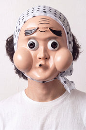 Hyottoko-Maske von einem Model vor hellem Hintergrund getragen — komisches Gesicht mit runden Wangen, schief gespitztem Mund und blauem Kopftuch