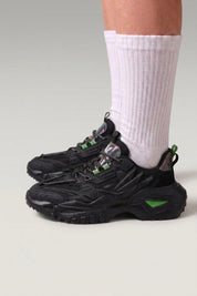 Chunky-Sneaker 'Shinshiro' in Schwarz — gerippte Linien an den Seiten, gesprenkelte Sohle, am Fuß mit weißen Socken vor grauem Studiohintergrund