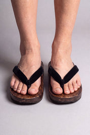 Japanische Flip Flops in uni schwarz, barfuß von vorn getragen — massive braune Holzsohle und gepolsterter Riemen zwischen den Zehen