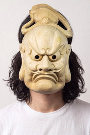 Bugaku-Maske von einem Model vor hellem Hintergrund getragen — strenges Gesicht aus hellem Holz mit gebogener Kopfbedeckung und gerunzelten Brauen