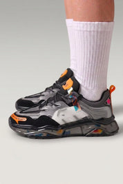 Chunky-Sneaker 'Hekinan' in Schwarz — transparente Sohle voller bunter Splitter, am Fuß mit weißen Socken vor grauem Studiohintergrund