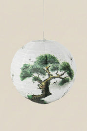 Japanische Laterne in Baum — weiße Papierkugellaterne mit knorriger Kiefer in grüner und schwarzer Tusche, an einer Schnur vor beigem Hintergrund