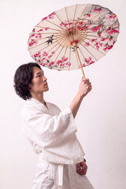 Japanischer Wagasa 'Nomikomu' geöffnet, von einem Model im weißen Kimono gehalten — cremefarbene Bespannung voller rosa Kirschblüten