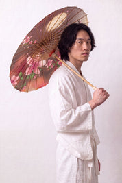 Japanischer Wagasa 'Chisana' geöffnet, von einem Model im weißen Kimono gehalten — kleine Bespannung im Braun-Rosa-Verlauf mit Kirschblüten