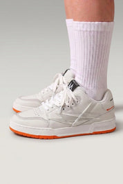 Chunky-Sneaker 'Inazuma' in Orange — weiß-graues Obermaterial auf kräftig einfarbiger Sohle, am Fuß mit weißen Socken vor grauem Studiohintergrund