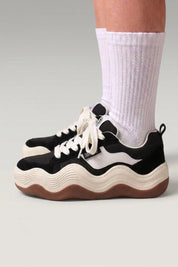 Plateau-Sneaker 'Nami' in Schwarz — Plateausohle als zweifarbige Wellenlinie geschnitten, am Fuß mit weißen Socken vor grauem Studiohintergrund