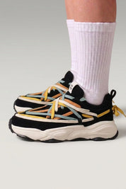 Chunky-Sneaker 'Fujioka' in Schwarz — doppelte bunte Schnürung, dicke cremefarbene Sohle, am Fuß mit weißen Socken vor grauem Studiohintergrund