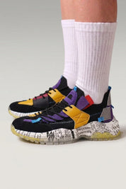 Chunky-Sneaker 'Iwakura' in Schwarz & Purple — Zeitungsdruck, farbige Riemen, getönte Sohle, am Fuß mit weißen Socken vor grauem Studiohintergrund
