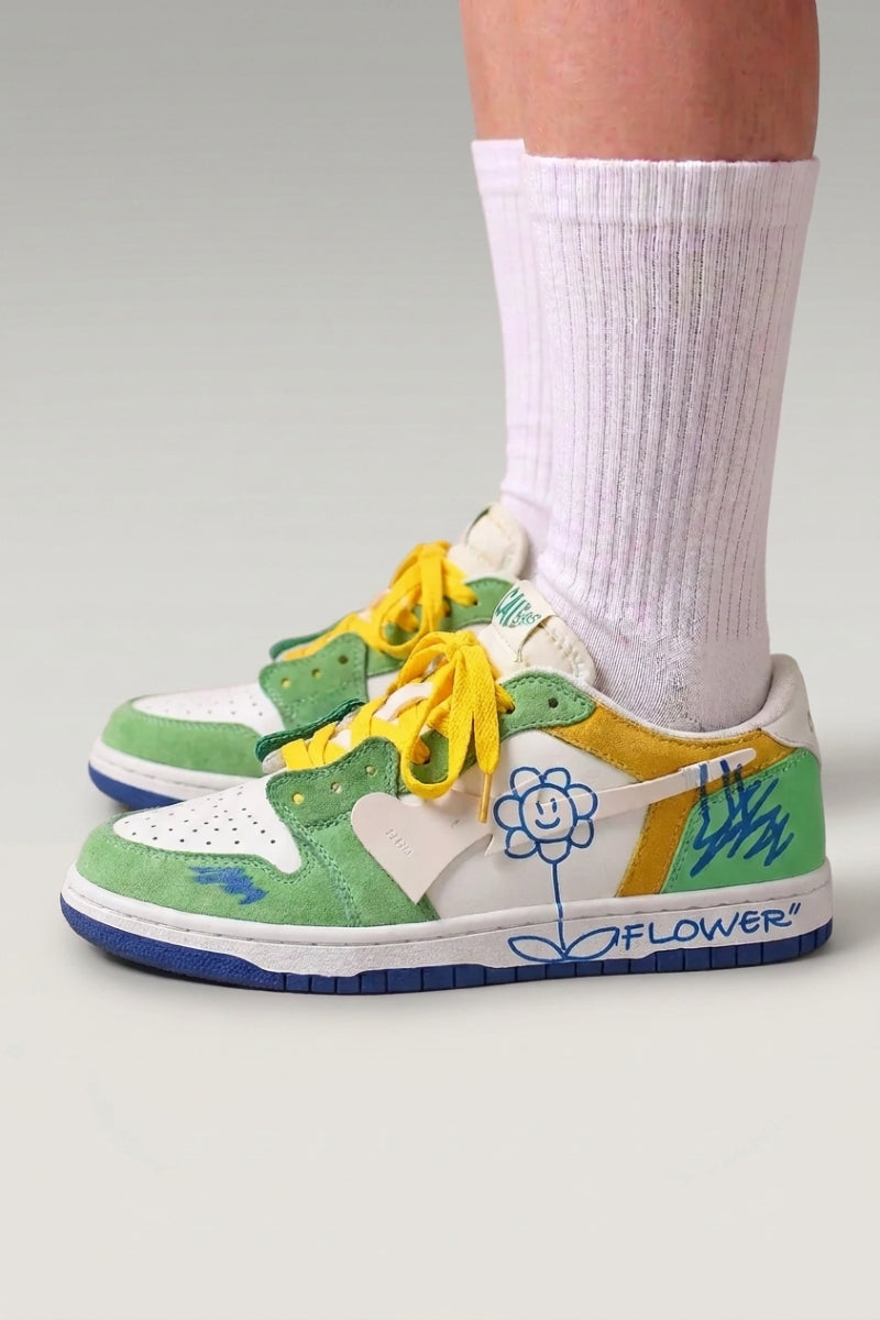 Low-Top-Sneaker 'Hanabi' in Grün — bunte Patches, aufgenähte Blume an der Ferse, breite Schnürsenkel, am Fuß mit weißen Socken vor grauem Studiohintergrund