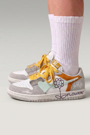 Low-Top-Sneaker 'Hanabi' in Weiß — bunte Patches, aufgenähte Blume an der Ferse, breite Schnürsenkel, am Fuß mit weißen Socken vor grauem Studiohintergrund
