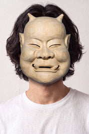 Beige Noh-Maske von einem Model vor hellem Hintergrund getragen — lächelndes altes Gesicht mit zusammengekniffenen Augen, sandfarben und schlicht modelliert
