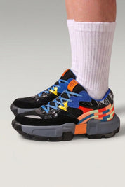 Chunky-Sneaker 'Tsuruga' in Schwarz — kontrastierende blaue und orange Panels, Gummisohle, am Fuß mit weißen Socken vor grauem Studiohintergrund