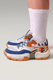 Chunky-Sneaker 'Tsuruga' in Weiß — kontrastierende blaue und orange Panels, Gummisohle, am Fuß mit weißen Socken vor grauem Studiohintergrund