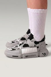 Low-Top-Sneaker 'Kazuno' in Grau — Utility-Riemen, Metallösen, segmentierte Sohle, am Fuß mit weißen Socken vor grauem Studiohintergrund