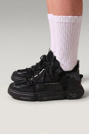 Low-Top-Sneaker 'Kazuno' in Schwarz — Utility-Riemen, Metallösen, segmentierte Sohle, am Fuß mit weißen Socken vor grauem Studiohintergrund